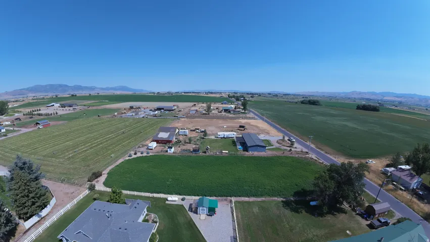 Expansive rural property featuring multiple outbuildings, agricultural fields, a paved road, and distant mountains under a clear sky