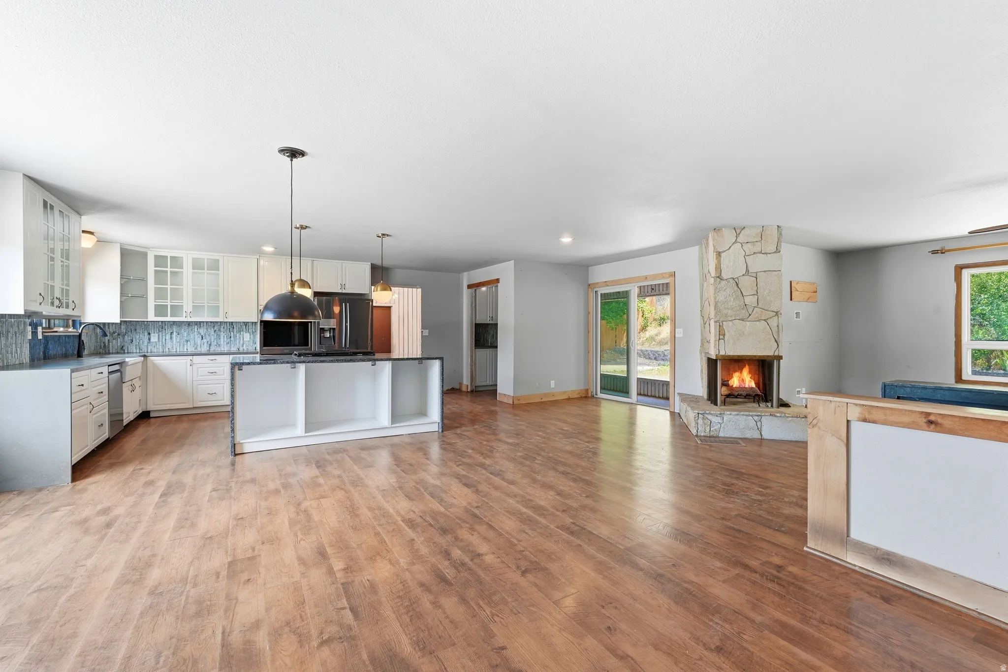 Open-concept living area featuring wood-finish flooring, a stone-clad fireplace, a full kitchen with an island, white cabinetry, and a tile backsplash, and a sliding glass door