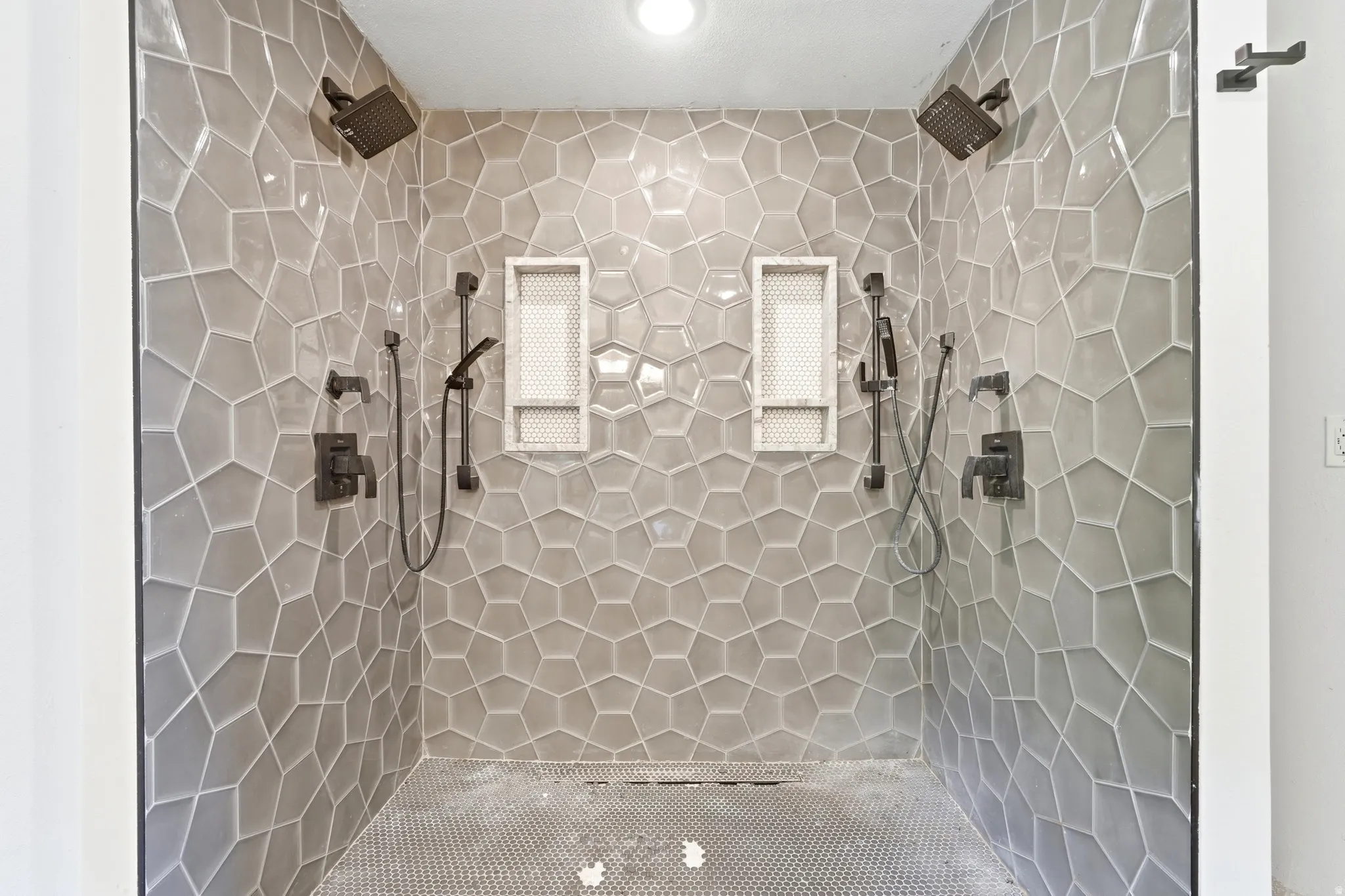 Walk-in shower featuring geometric tiled walls, dual overhead rain showerheads, two handheld shower wands, integrated shelving niches, and dark bronze fixtures