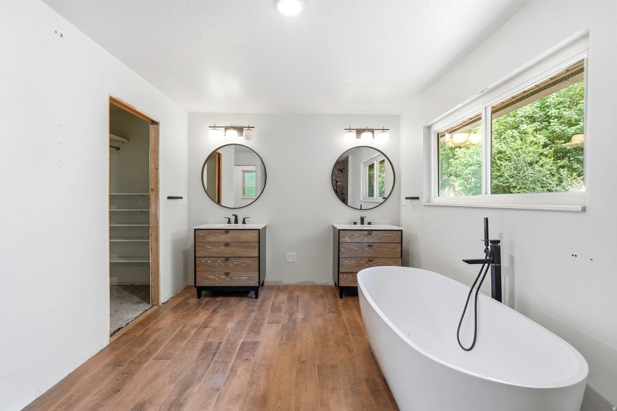 Bathroom featuring a freestanding white soaking tub with a matte black floor-mounted faucet, wood-finish flooring, dual vanity stations with integrated sinks and round mirrors, and a wide window