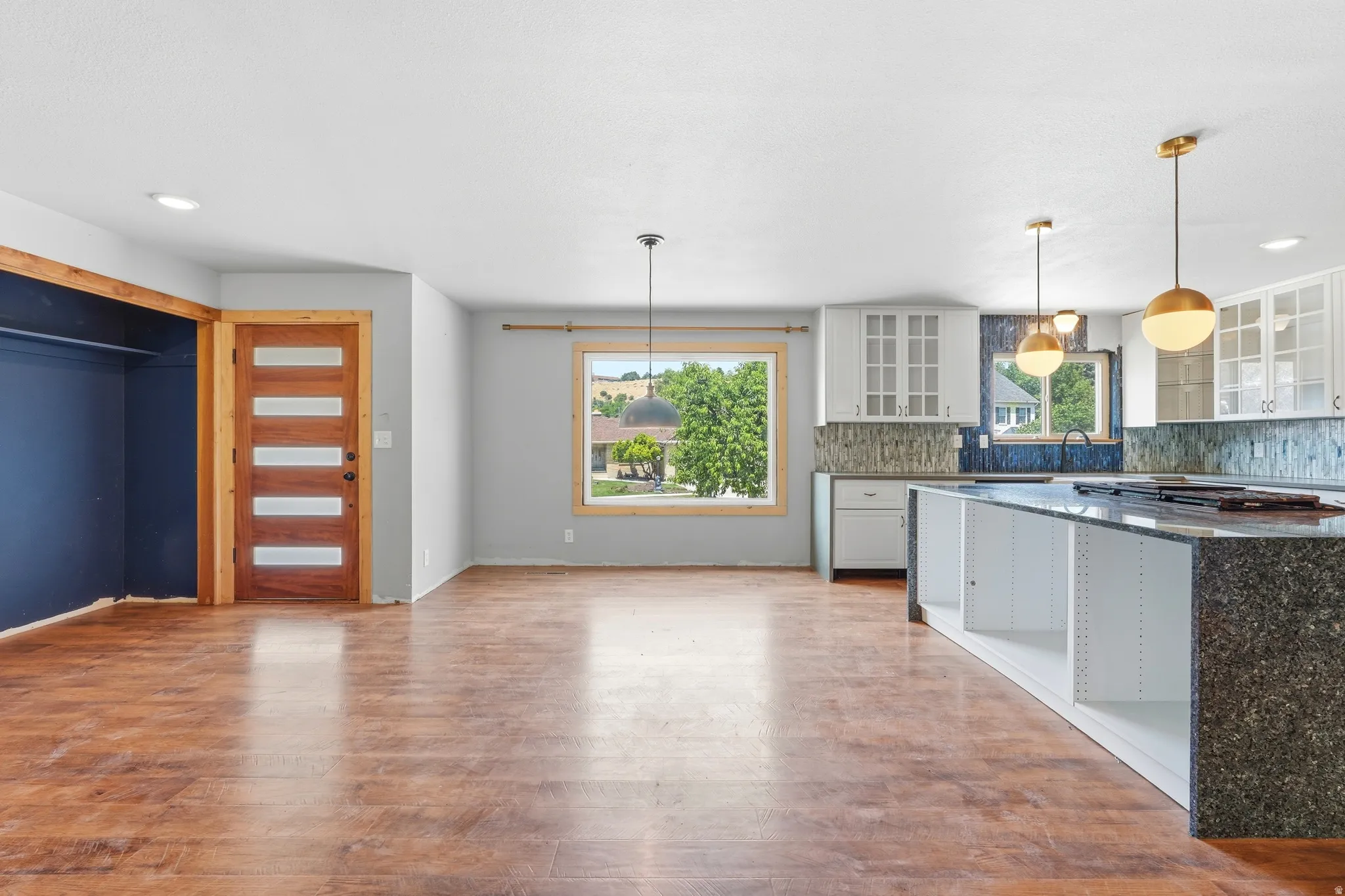 Open-concept living area featuring wood-finish flooring, a modern kitchen island with integrated cooktop, white shaker-style cabinetry, and contemporary pendant lighting