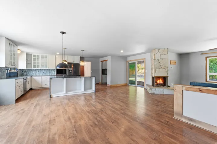 Open-concept living area featuring wood-finish flooring, a stone-clad fireplace, a full kitchen with an island, white cabinetry, and a tile backsplash, and a sliding glass door