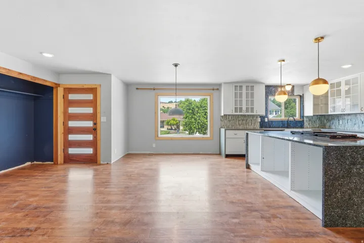 Open-concept living area featuring wood-finish flooring, a modern kitchen island with integrated cooktop, white shaker-style cabinetry, and contemporary pendant lighting