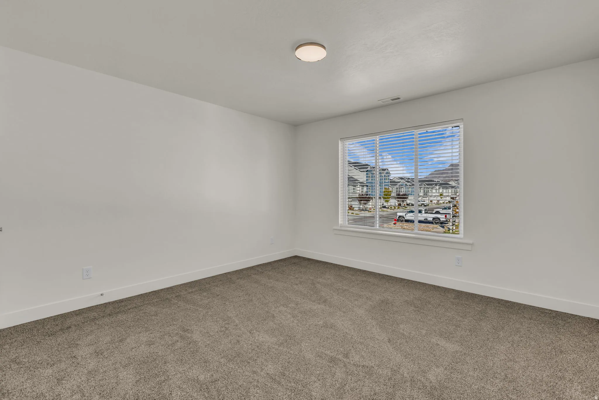 Neutral white walls complement the gray carpet flooring in this room featuring a window with horizontal blinds and a flush-mount ceiling light fixture