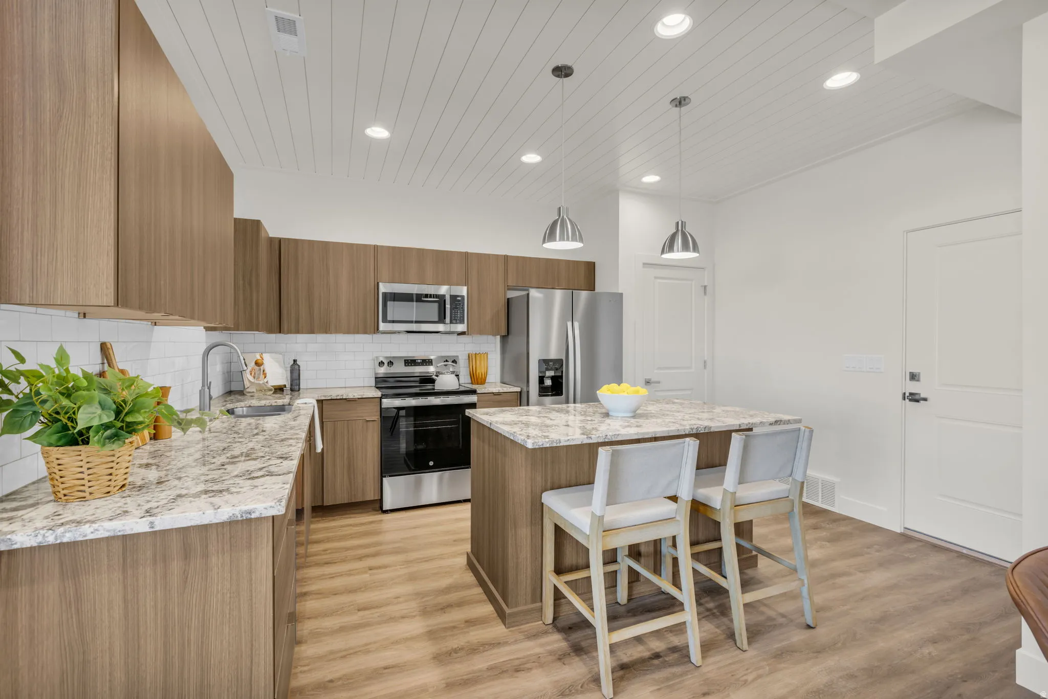 Contemporary kitchen featuring wood-finish cabinetry, light-toned countertops, a full suite of stainless steel appliances, a subway tile backsplash, and recessed and pendant lighting