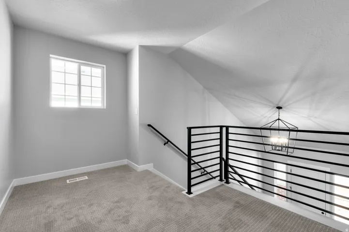 Upper-level landing featuring a single window, textured ceiling, light gray carpet, white baseboards, and a black horizontal bar railing with a corresponding handrail
