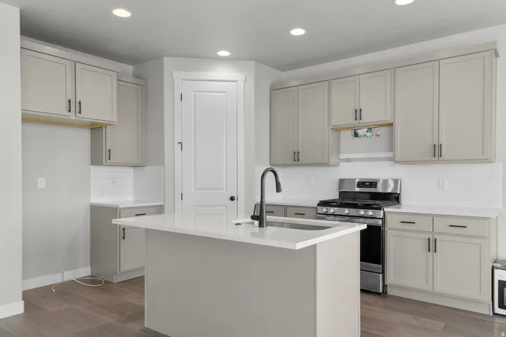 Kitchen featuring an island with an undermount sink and matte black faucet, light gray cabinetry with black hardware, stainless steel range, white subway tile backsplash, and wood-finish flooring