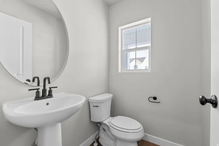 Pedestal sink with matte black faucet, round wall mirror, and toilet with a window above