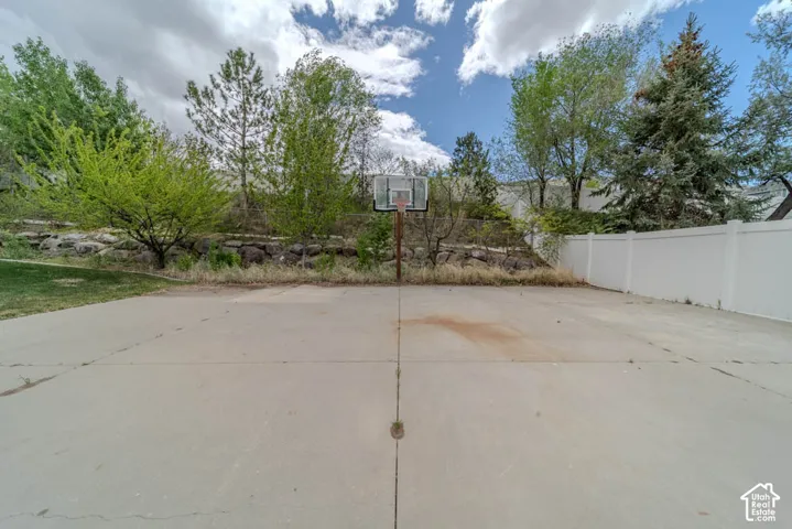 Concrete sports court featuring a basketball hoop, established landscaping, and a white privacy fence