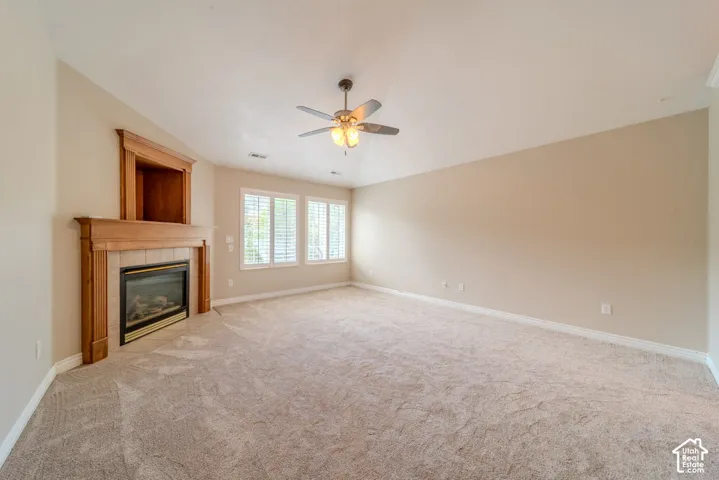 Spacious room featuring a fireplace with a wood-finish mantel and tile surround, built-in shelving, carpet flooring, a ceiling fan, and multiple windows with plantation shutters