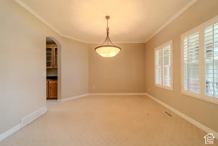 Neutral-toned room featuring a central chandelier, two windows with white plantation shutters, crown molding, and light-colored carpeting