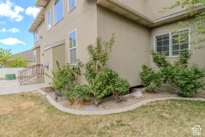Stucco exterior featuring multiple windows with louvered blinds, a concrete patio, and a landscaped garden bed with mature shrubs