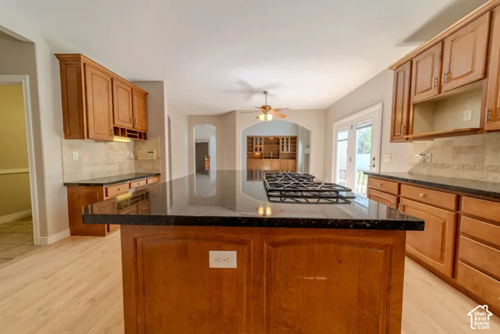Spacious kitchen featuring a large island with an integrated gas cooktop, rich wood-finish cabinetry, dark countertops, a tiled backsplash, and light wood-finish flooring