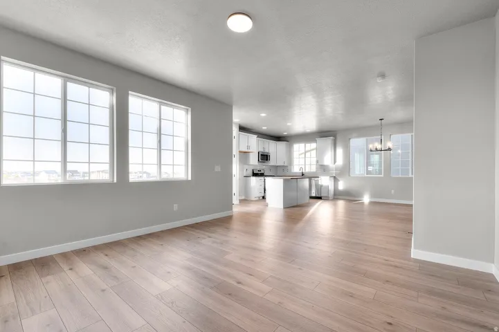 Open-concept living space featuring wood-finish flooring, ample natural light from multiple large windows, a central kitchen island, white cabinetry, and stainless steel appliances