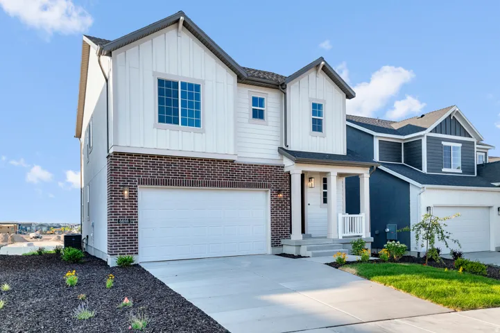 Modern townhome facade featuring white vertical siding, brick accents, a covered entry porch with columns, a two-car garage, and established landscaping