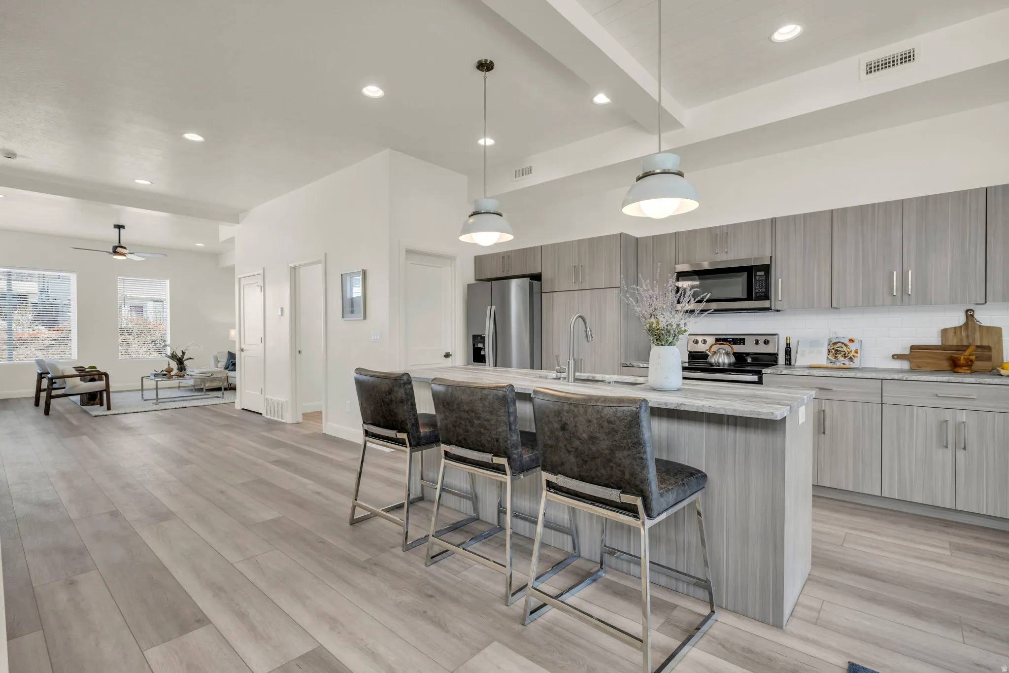 Open-concept kitchen and living area featuring grey wood-finish flooring, a kitchen island with pendant lighting, stainless steel appliances, and abundant natural light from multiple windows