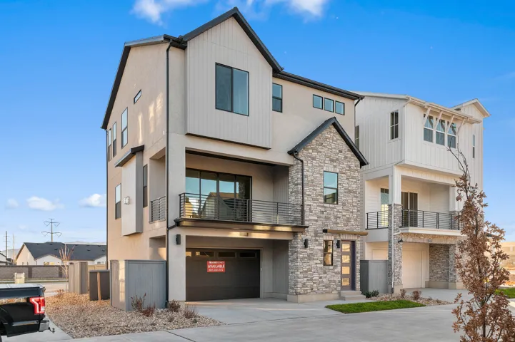Contemporary townhome featuring a stone facade, a dark garage door, a private balcony with black railings, and a prominent front entrance with a modern door