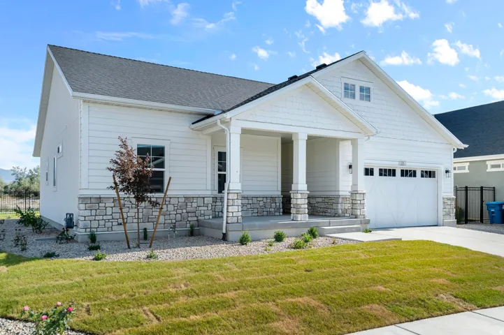 Contemporary exterior featuring white siding, a stone veneer foundation, a covered porch with stone-clad columns, a two-car garage, and a manicured lawn