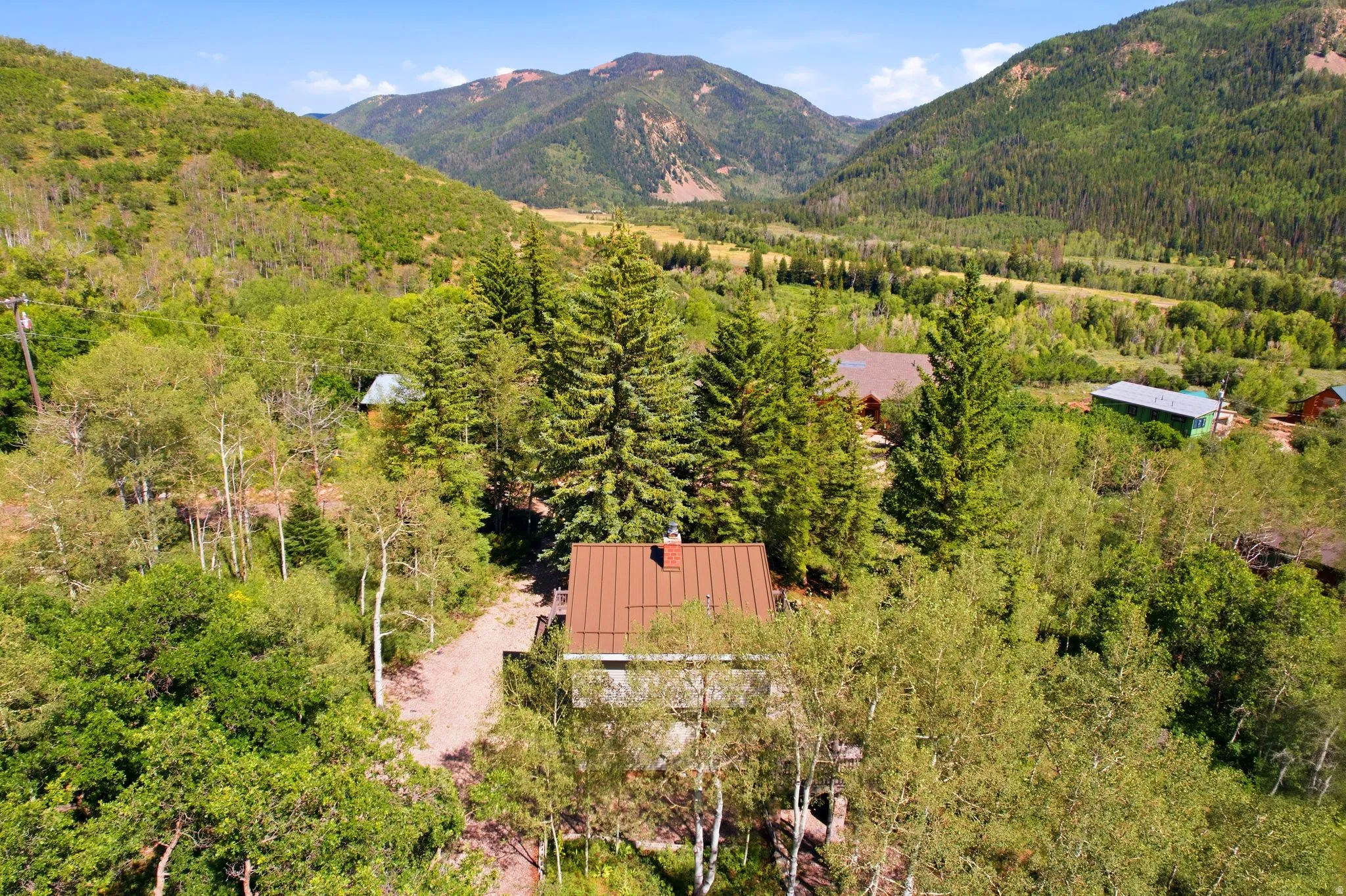 Mountainous landscape featuring a dwelling with a brown pitched roof, surrounded by numerous mature trees and a gravel pathway