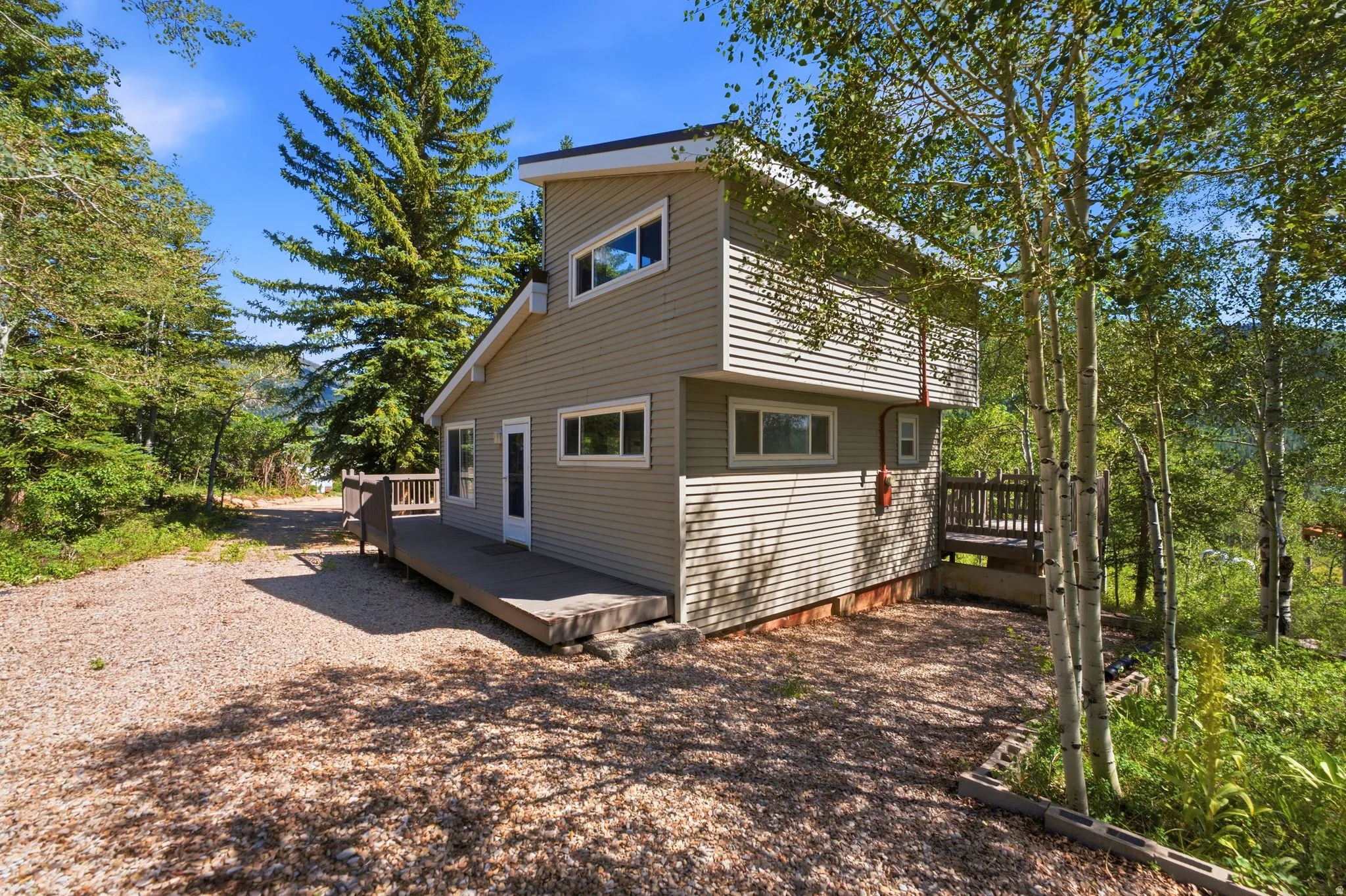 Two-story home with horizontal siding and multiple windows, featuring a gravel driveway and surrounding mature trees