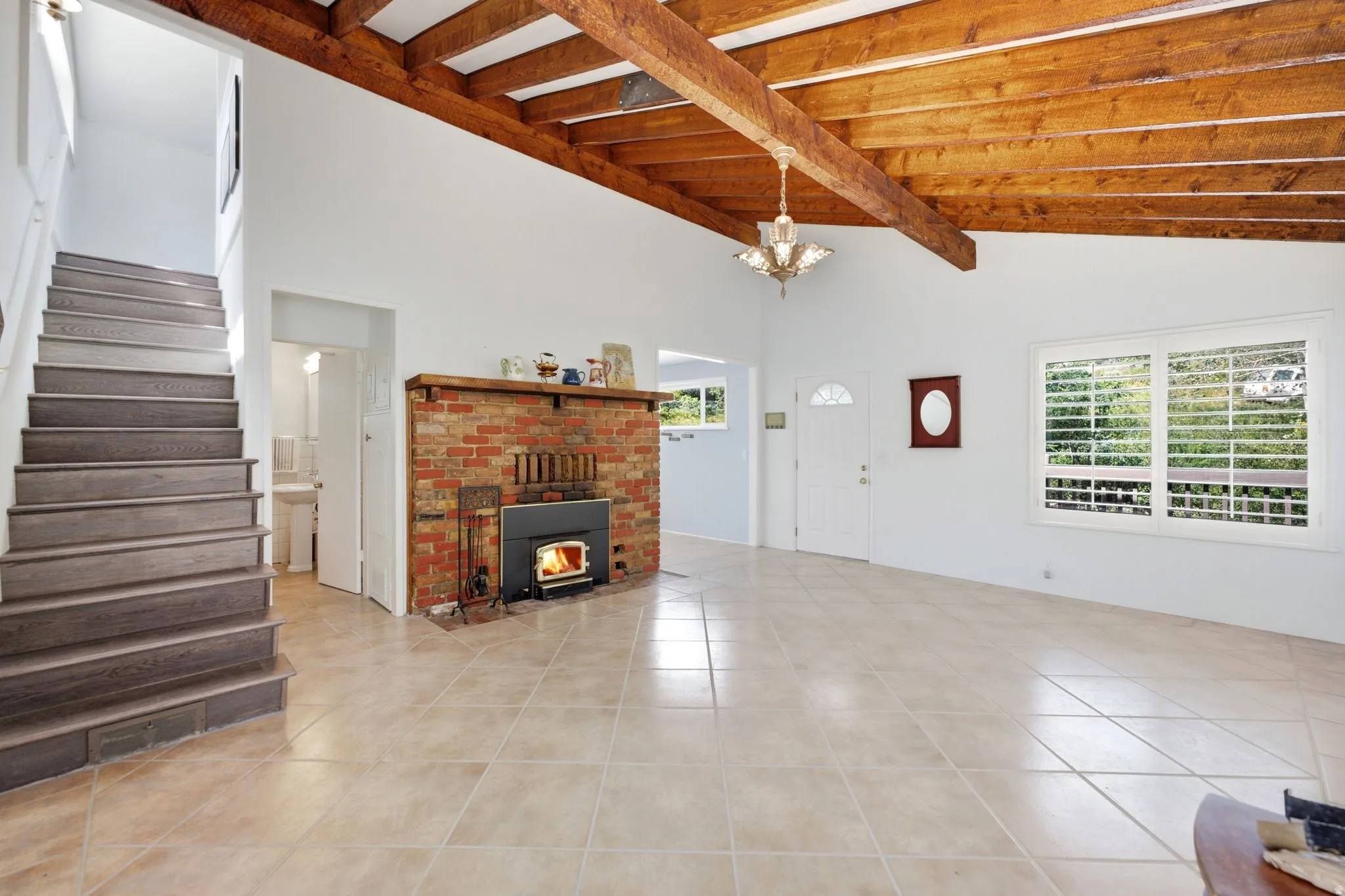 Spacious living area featuring a vaulted ceiling with exposed wood beams, a brick fireplace with a wood-burning insert, an open-tread staircase with wood-finish risers, large windows with interior shutters, and neutral-toned tile flooring