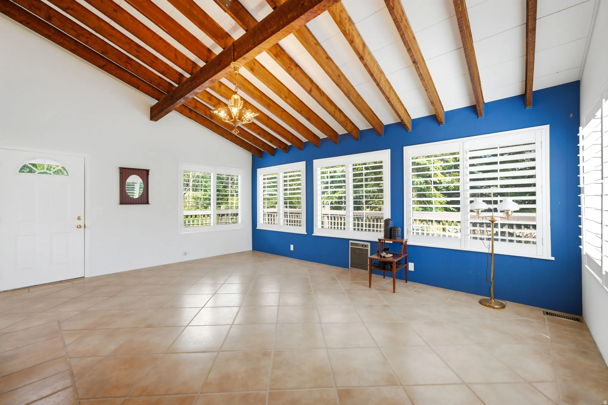 Vaulted exposed beam ceiling featuring a hanging chandelier, ceramic tile flooring, multiple windows with white plantation shutters, and a white entry door with an arched glass insert
