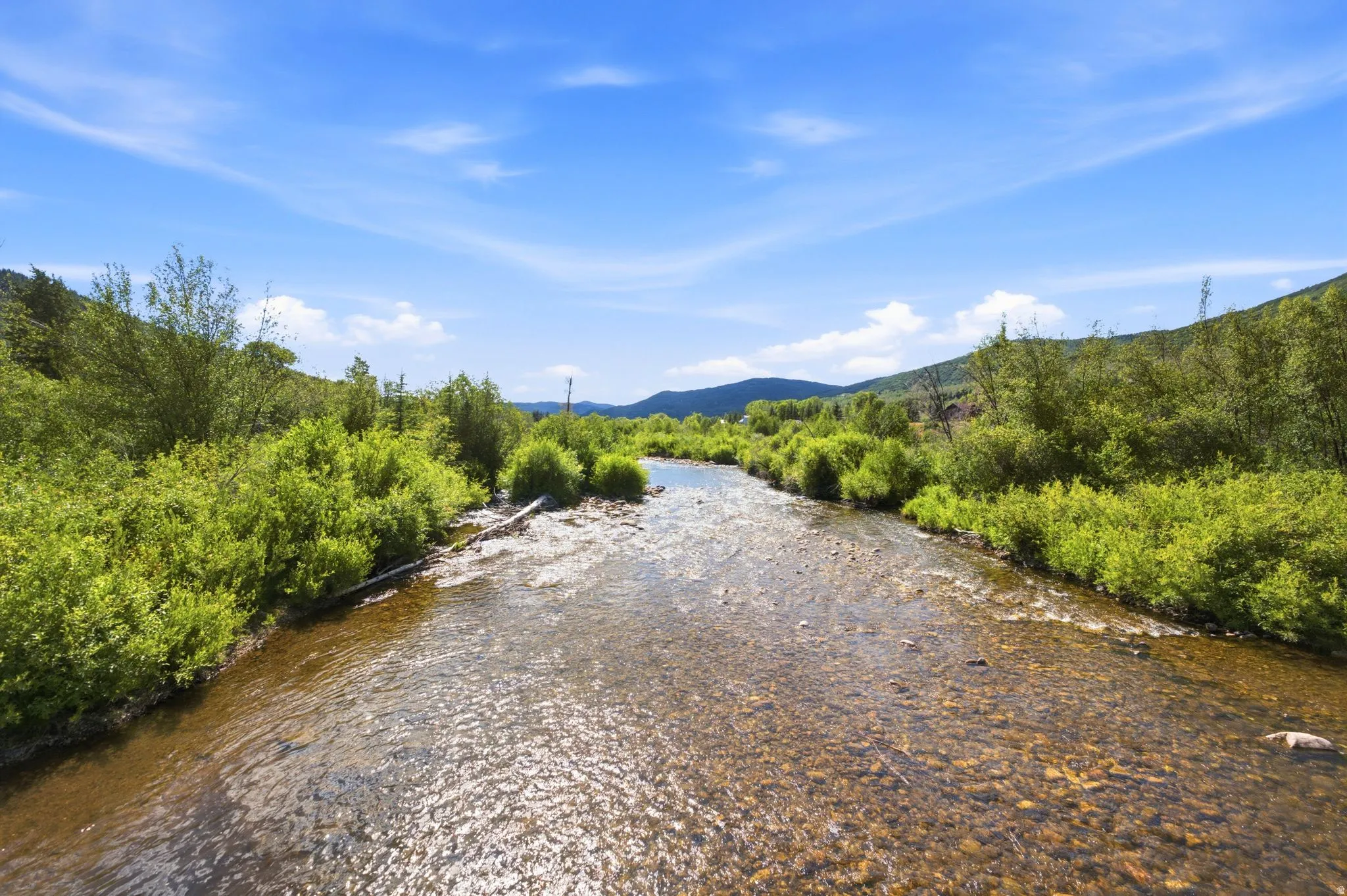Clear river with a rocky bed, flanked by lush green foliage and distant hills under a bright blue sky