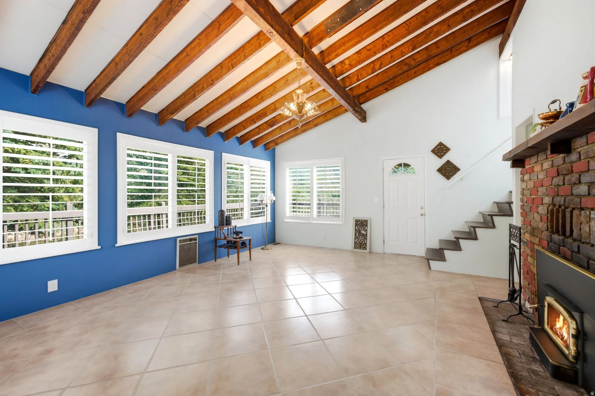 Vaulted ceiling with exposed wood beams, tile flooring, multiple windows with plantation shutters, brick fireplace with wood mantel, and an interior staircase