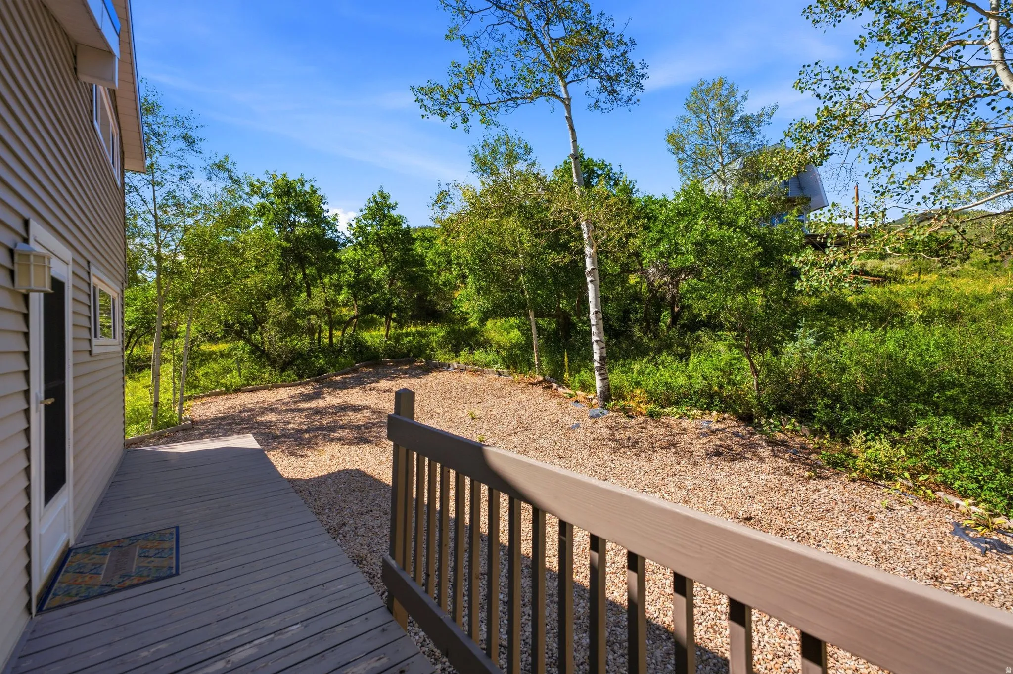 Expansive outdoor deck with wood-finish railing, overlooking a natural landscape with mature trees and gravel ground cover