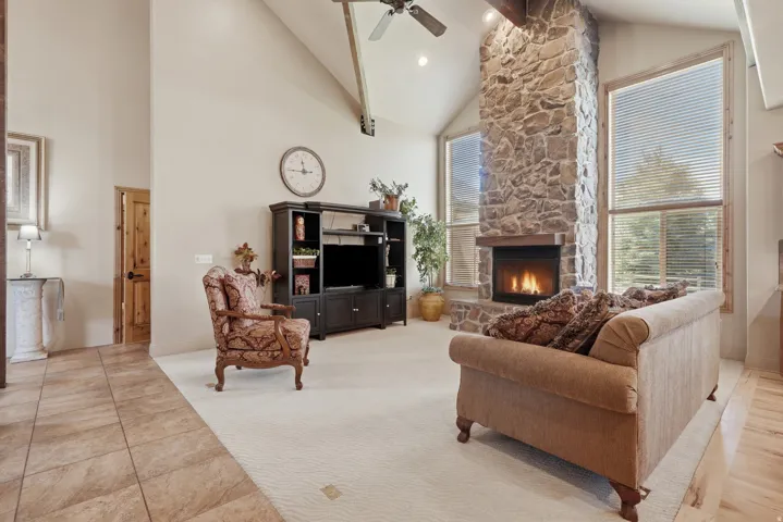 Vaulted ceiling living area featuring a floor-to-ceiling stone fireplace with a wood mantle, large windows with blinds, and light-toned wall finishes