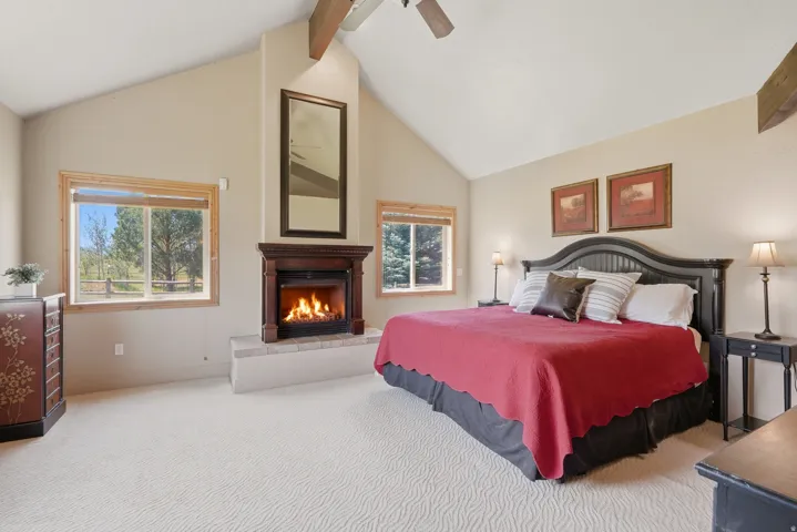 Vaulted ceiling room featuring a wood-mantled fireplace, natural wood-trimmed windows, a ceiling fan, and light-toned patterned carpeting