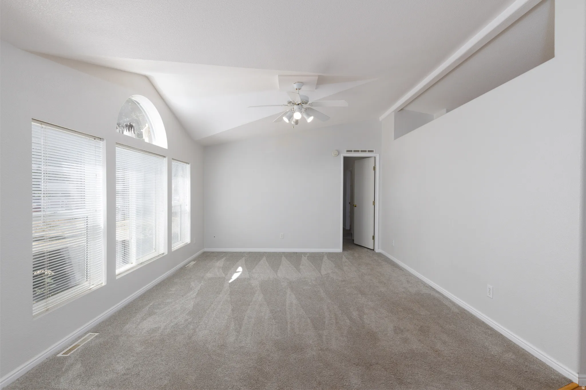 Spacious room featuring vaulted ceilings, a prominent arched window, two additional rectangular windows with blinds, light gray wall paint, and a ceiling fan with integrated lighting