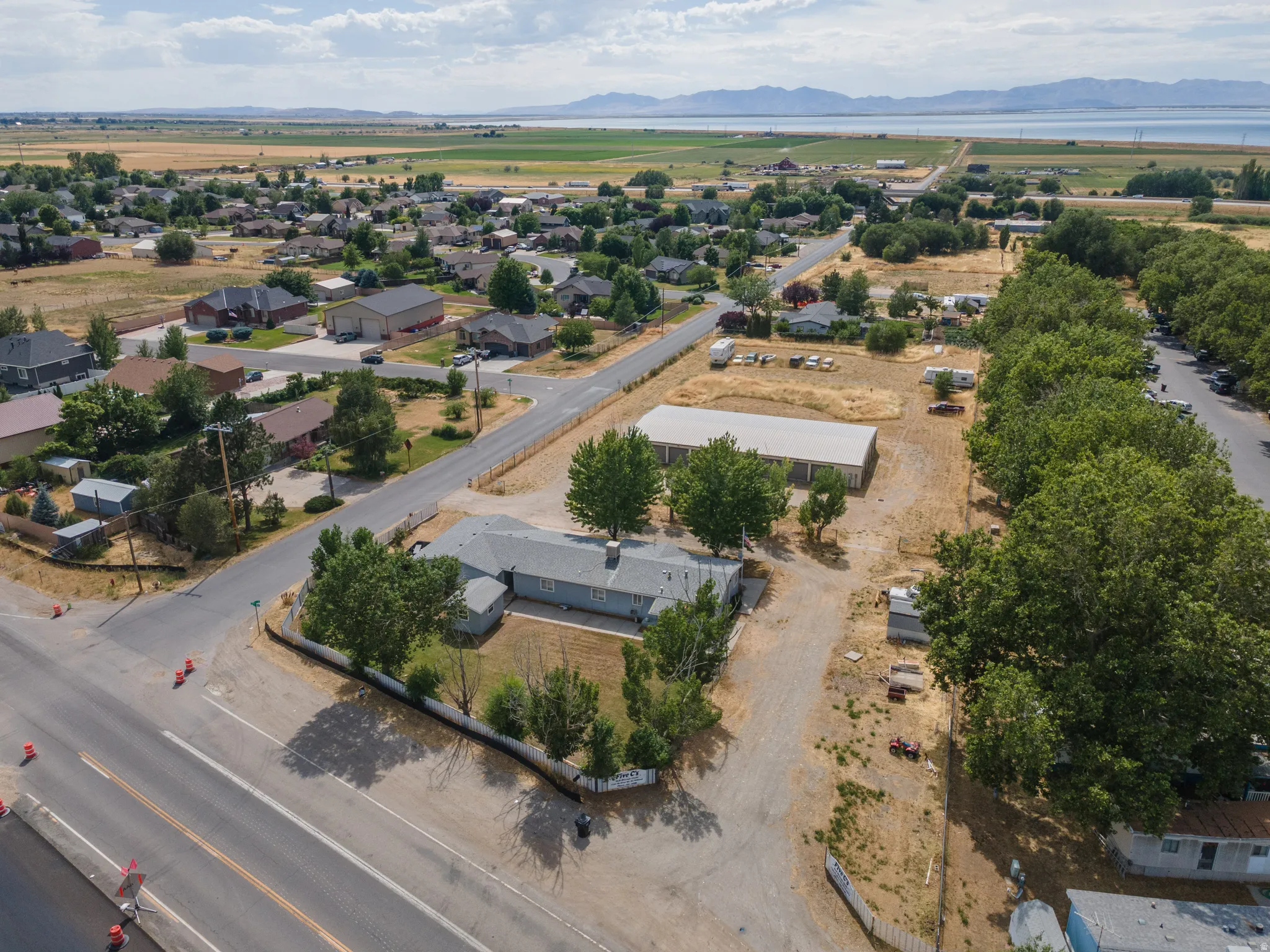 Expansive lot featuring a single-story residence with a light gray exterior, a large outbuilding, mature perimeter trees, and a distant mountain and water backdrop