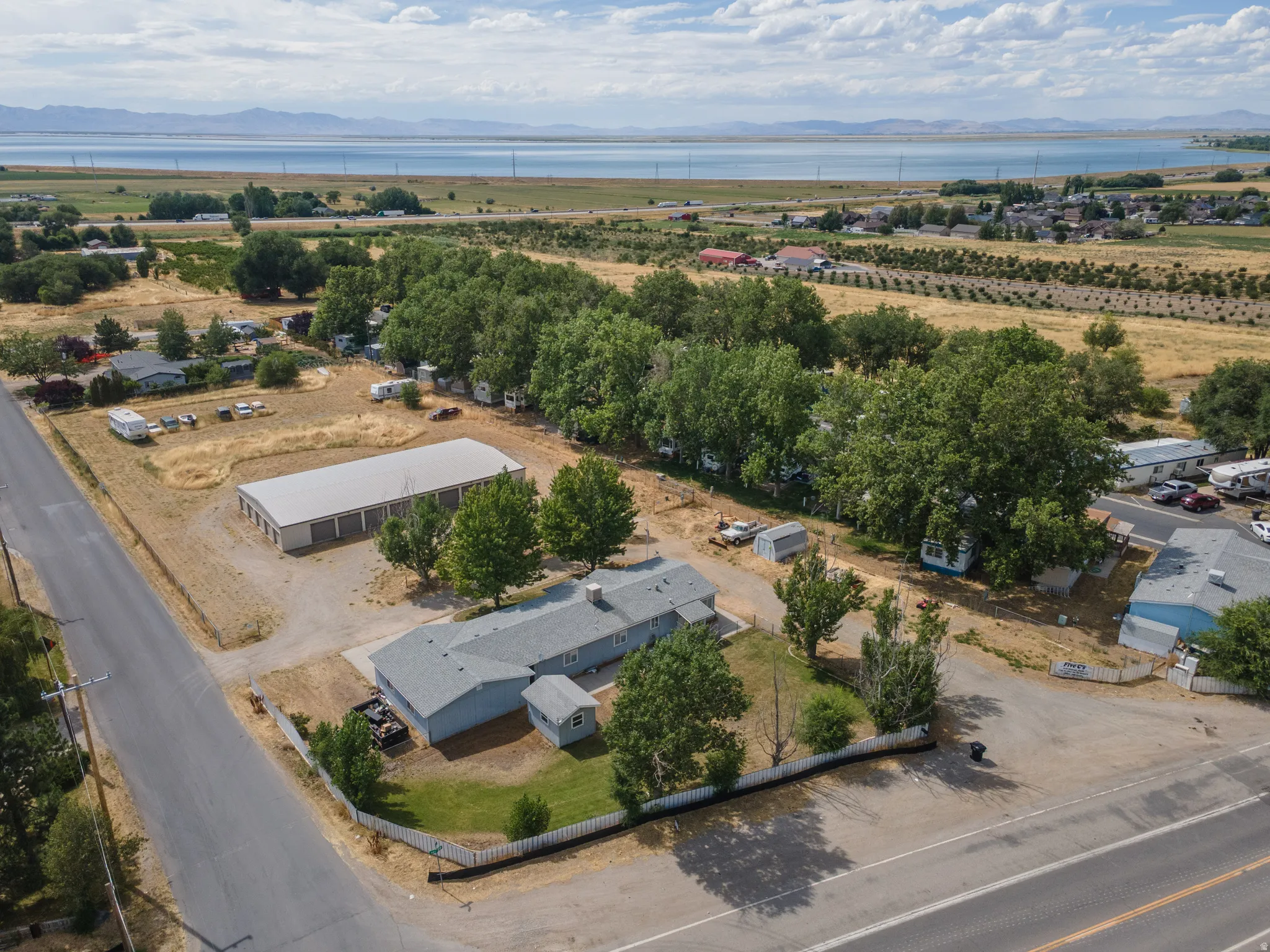 Expansive property featuring a single-story residence with a gray roof, a large multi-bay storage building, mature trees, and a distant water body