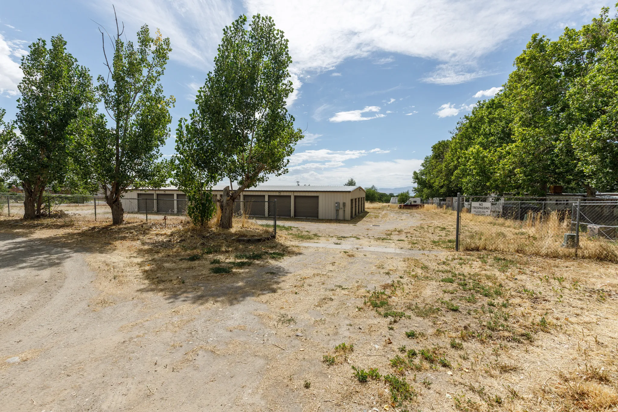 Multiple storage buildings with roll-up doors, chain-link fencing, and mature trees on a gravel lot
