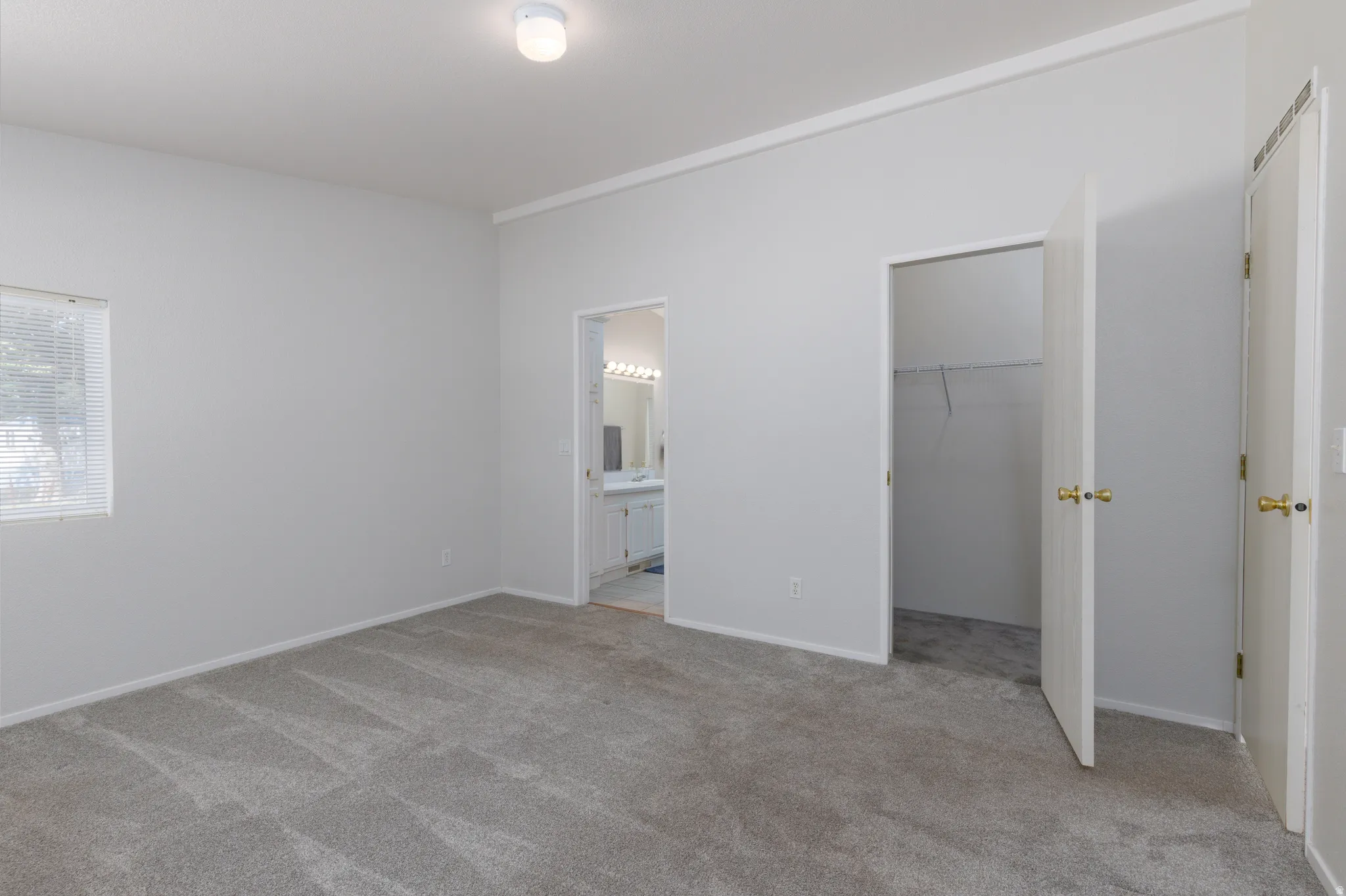 Carpeted room featuring a window with blinds, a ceiling-mounted light fixture, a closet with bi-fold doors, and an entryway to a bathroom with a dual vanity