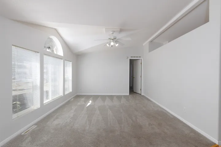 Spacious room featuring vaulted ceilings, a prominent arched window, two additional rectangular windows with blinds, light gray wall paint, and a ceiling fan with integrated lighting