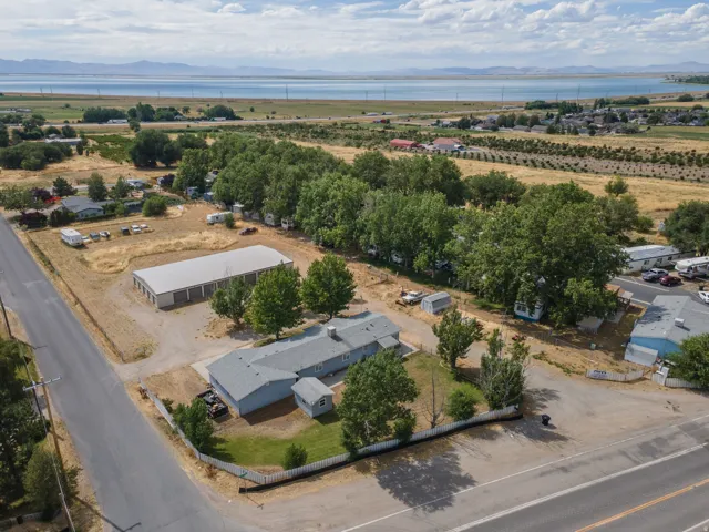Expansive property featuring a single-story residence with a gray roof, a large multi-bay storage building, mature trees, and a distant water body