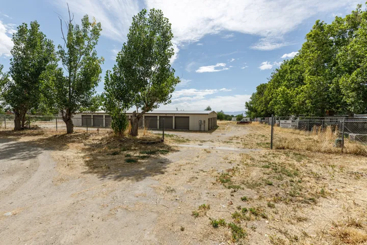 Multiple storage buildings with roll-up doors, chain-link fencing, and mature trees on a gravel lot