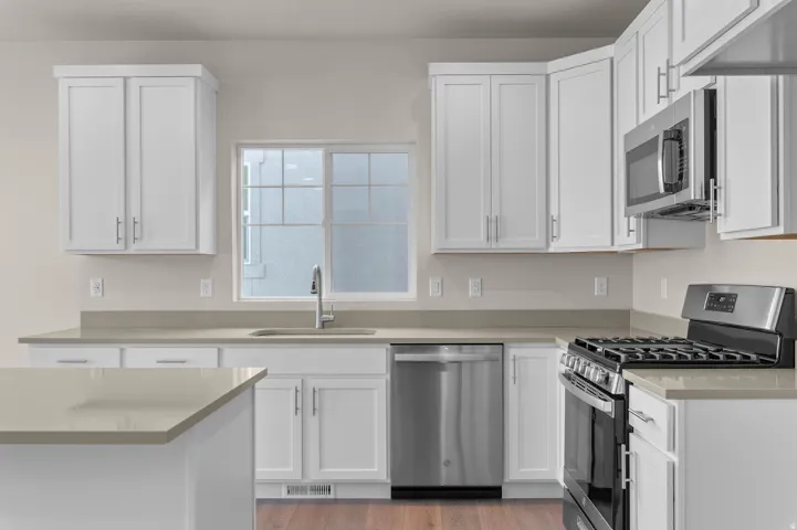 Bright kitchen featuring white shaker cabinetry, stainless steel appliances, a corner window above the sink, light-toned countertops, and wood-finish flooring
