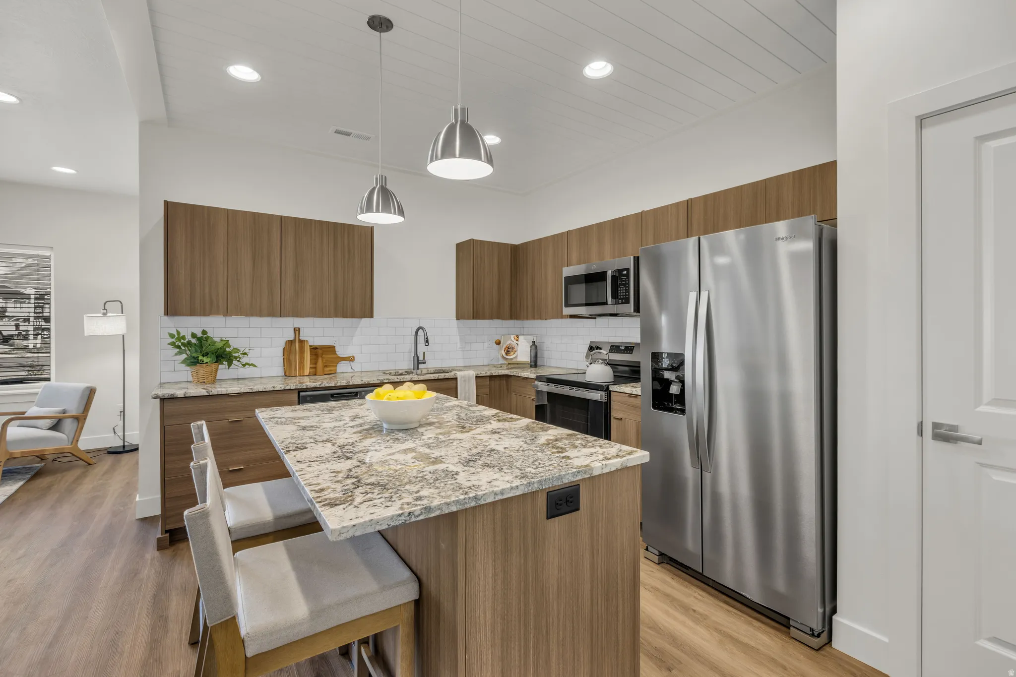 Open-concept kitchen featuring an island with counter seating, stainless steel appliances, wood-finish cabinetry, and a subway tile backsplash