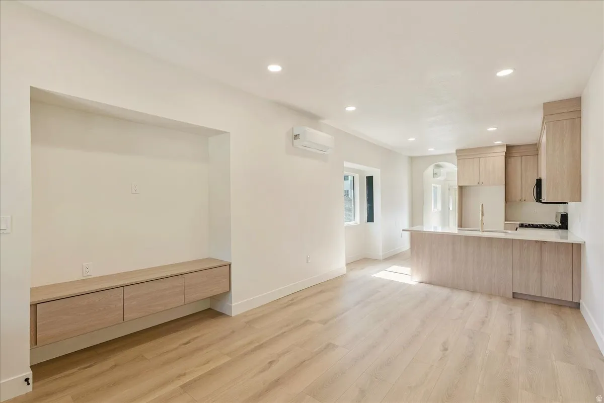 Open-concept living space with light wood-finish flooring, recessed lighting, a built-in media niche with floating cabinetry, and a modern kitchen featuring light wood cabinetry, a peninsula with stone-finish countertop, and a built-in cooktop with oven
