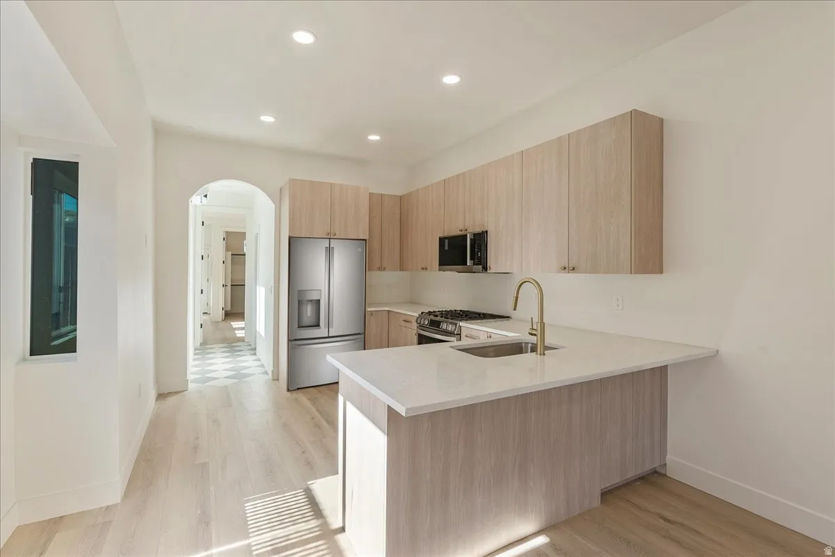 Modern kitchen featuring light wood-finish cabinetry, stainless steel appliances, a white countertop, and a brass-finish faucet