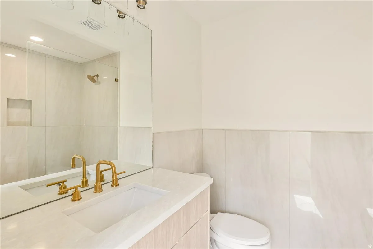 Bathroom featuring a large vanity mirror, gold-finish fixtures, an integrated sink, a glass-enclosed shower, and light-toned wall tiling