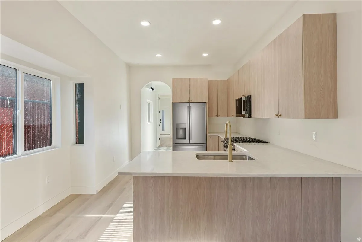 Modern kitchen featuring light wood-finish cabinetry, light-toned countertops, an undermount sink with a brass-finish faucet, and stainless steel appliances