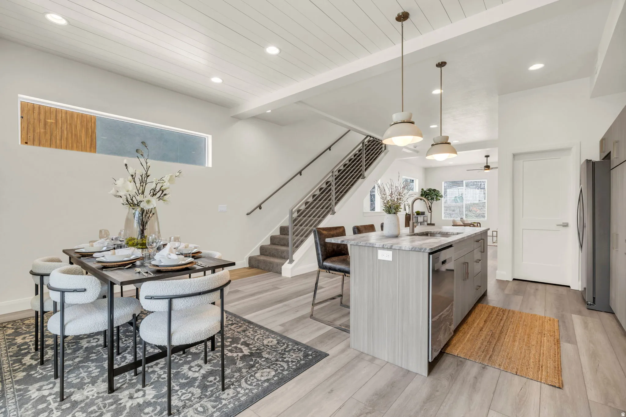 Open-concept living space with wood-finish flooring, a contemporary kitchen island with a stone countertop, modern pendant lighting, and a staircase with metal railings