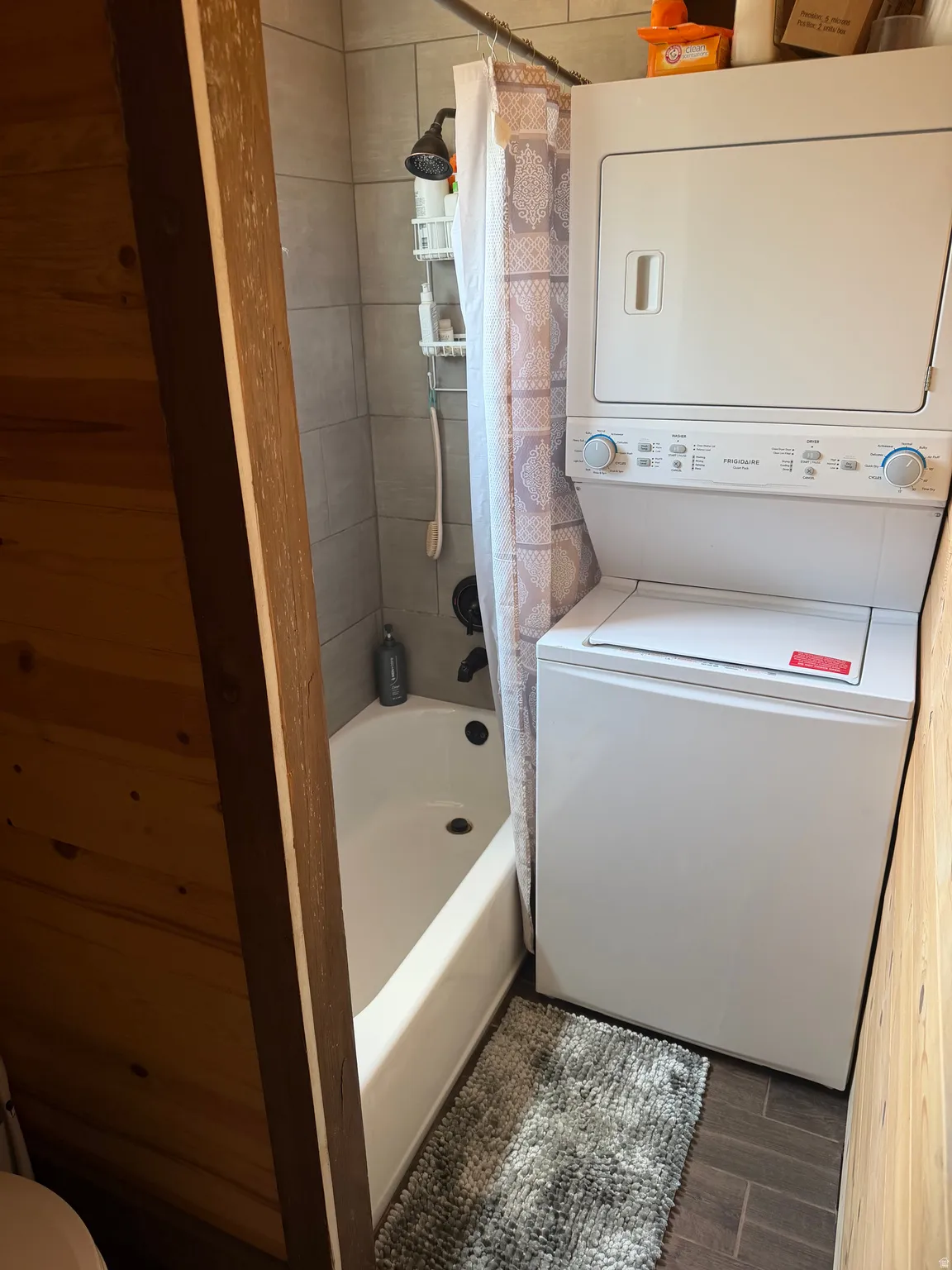 Vertical stacked washer and dryer unit beside a white bathtub with a tiled shower surround and dark bronze fixtures