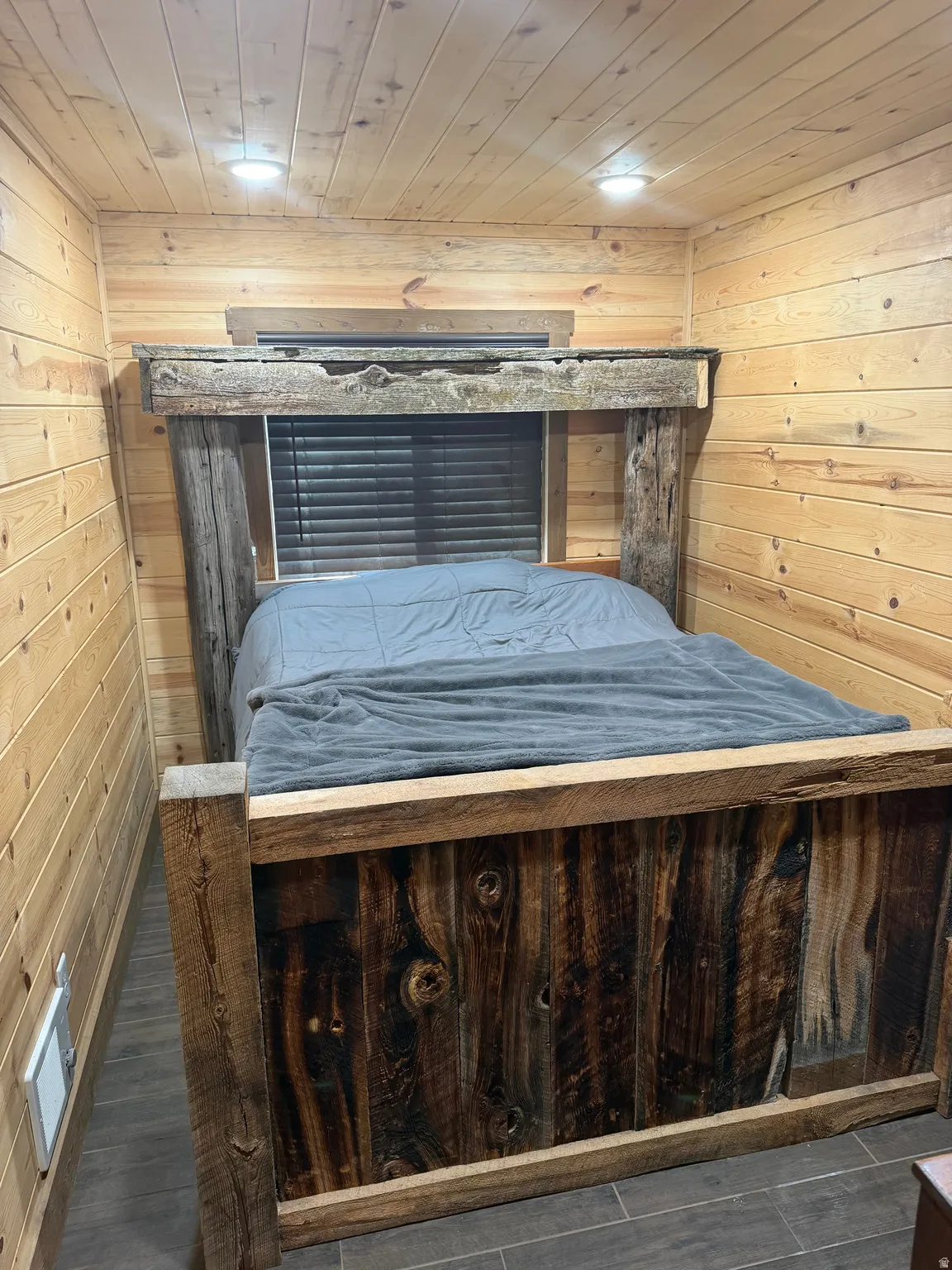 Reclaimed wood-style bed frame, wood-paneled walls and ceiling, recessed lighting, window with blinds, and tile flooring