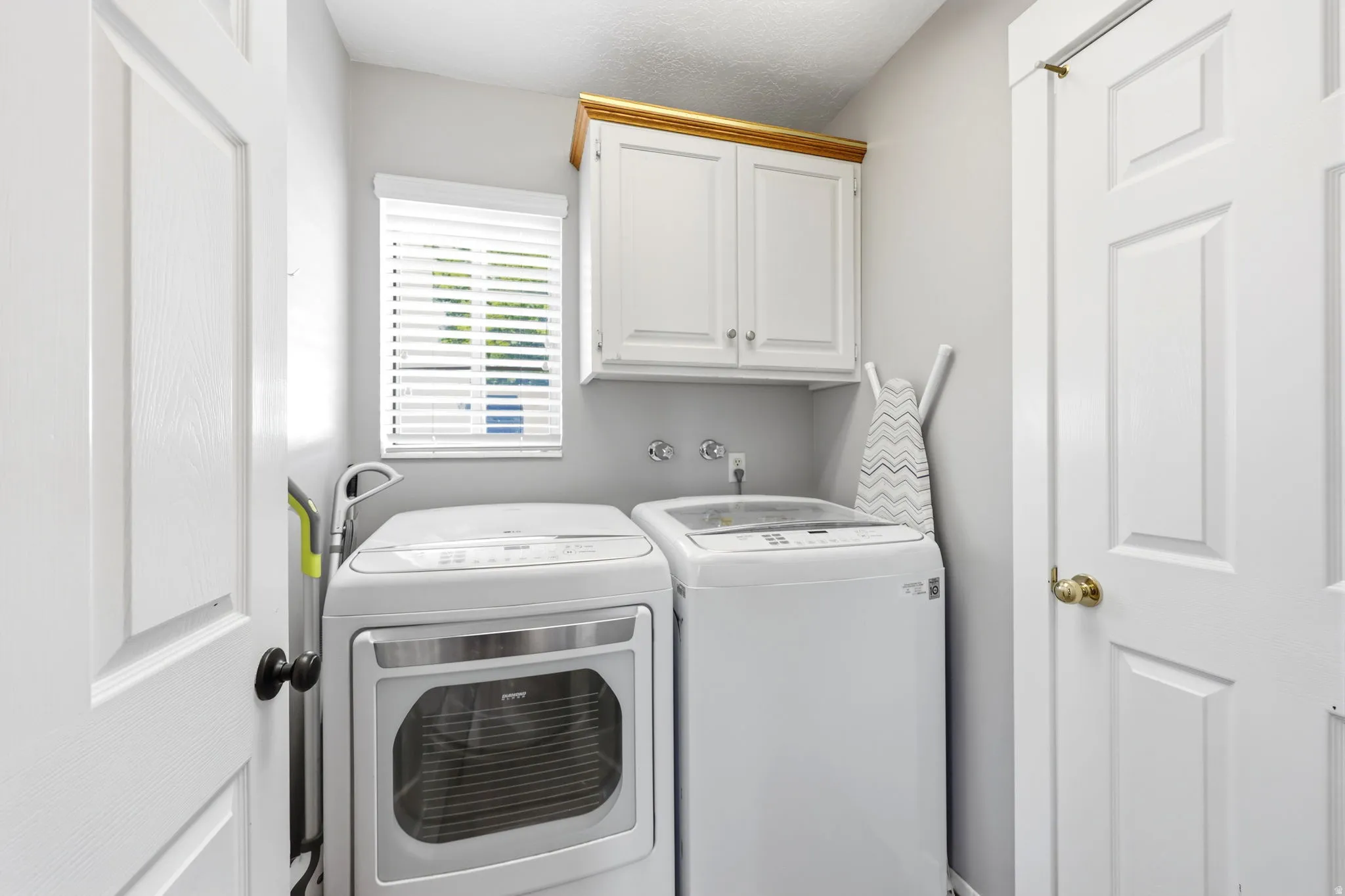 Dedicated laundry area featuring white shaker-style cabinetry with gold trim, a window with horizontal blinds, and paneled doors with black and brass hardware