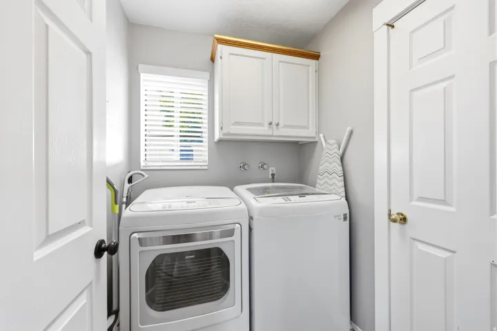 Dedicated laundry area featuring white shaker-style cabinetry with gold trim, a window with horizontal blinds, and paneled doors with black and brass hardware